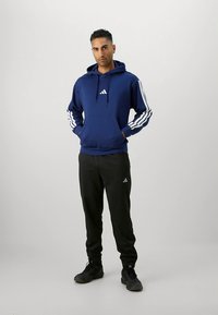 Μπλε σκούφο φούτερ με λευκές τρεις ρίγες στα μανίκια, μπροστινή τσέπη τύπου καγκουρό και λογότυπο. Συνδυασμένο με μαύρο παντελόνι και σκούρα αθλητικά παπούτσια.