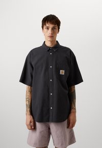 Camicia grigia a maniche corte con un motivo testurizzato, chiusura a bottoni sul davanti e taschino sul petto con un'etichetta del logo. Indossata con pantaloni corti rosa chiaro.