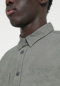 Πράσινο πουκάμισο με κουμπιά και μυτερό γιακά, με υφή υφάσματος, δύο μπροστινές τσέπες και γκρι κουμπιά, κοντινή λήψη στο λαιμό και τον ώμο ενός μοντέλου.