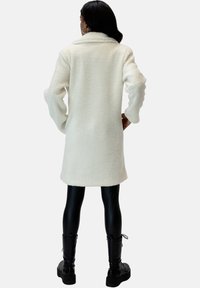 Hvid fleecefrakke med krave, lange ærmer og tekstureret finish. Sættes sammen med sorte leggings og snørebåndsankelstøvler.