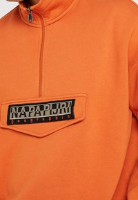 Orange fleece pullover with a high collar and half zipper. Features a black patch with "NAPAPIJRI geographic" in gray and red lettering.