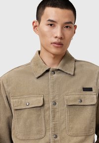 Beige kordjacka med två bröstfickor, krage och silverfärgade tryckknappar, som visar ribbad textur genom hela plagget.