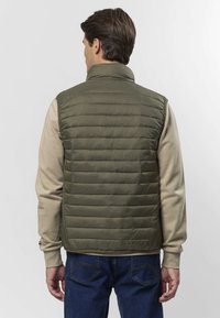 Vihreä toppaliivi vaakasuorin tikkausompelein, korkealla kauluksella, beige neuleen päällä. Klassiset farkut ja yksivärinen tausta täydentävät asun.