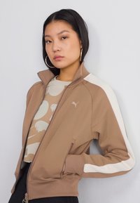 Brown zip-up jacket with white side accents, ribbed cuffs, and a fitted design. Worn over a light graphic t-shirt. Smooth fabric texture.