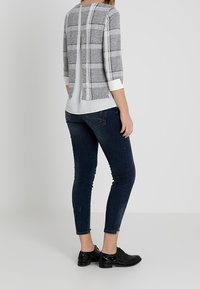 Haut à carreaux gris et blancs avec manches trois-quarts, superposé sur une chemise blanche ; assorti à un jean skinny foncé et des chaussures noires.