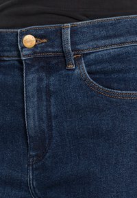 Pantalones vaqueros de denim azul oscuro con botón de cobre y costuras naranjas, mostrando el bolsillo delantero y el lazo del cinturón, combinados con una camiseta negra.