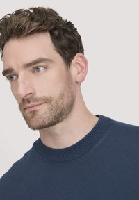 Blauer Strickpullover mit rundem Ausschnitt und strukturierten Rippendetails. Der Stoff wirkt weich mit subtiler Textur. Das Model zeigt einen ruhigen Ausdruck.