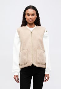 Vest din fleece bej cu fermoar, două buzunare frontale și detalii contrastante; purtat peste o cămașă albă și asortat cu pantaloni negri.