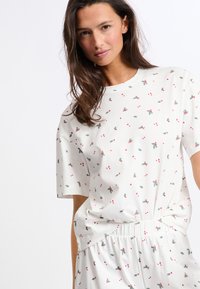 Camisa blanca de manga corta con cuello redondo, que presenta un pequeño patrón de flores y corazones en rojo y verde. Tejido suave y ligero.