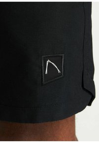 Pantaloni corti neri con un'etichetta logo cucita in un design quadrato. Una consistenza evidente e dettagli delle cuciture mettono in risalto la durata del tessuto.