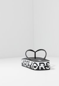 Musta flip-flop sydämenmuotoisella hihnalla ja teksturoidulla pohjalla. Varustettu rohkealla valkoisella logonauhalla välikannan ympärillä.