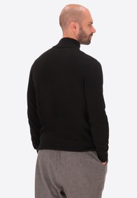 Maglione nero a maglia con colletto alto, slim fit e orlo a costine. Il modello indossa pantaloni grigi, in posa di profilo. Texture liscia, design minimal.