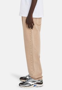 Ανοιχτό καφέ, άνετο τζιν με χαλαρή γραμμή, με πλαϊνή τσέπη και αντίθετες ραφές. Συνδυασμένο με μαύρα και ασημί αθλητικά παπούτσια.