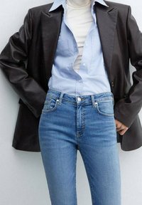 Blazer de cuero sintético negro sobre una camisa de botones azul claro y un cuello de tortuga blanco, combinado con jeans azules de cintura alta.