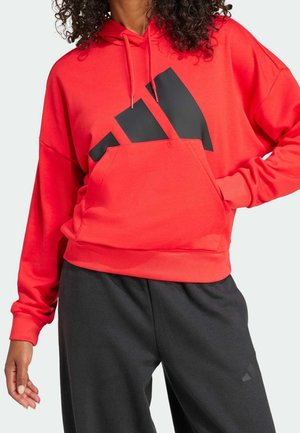 Persoon draagt een rode hoodie met zwart logo en trekkoorden, gecombineerd met losse zwarte broek, staand met één hand in de zak.