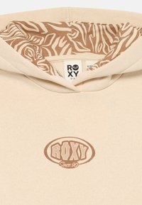 Beige hoodie med brun "Roxy Since 90" logotyp på framsidan och zebramönstrad tygfoder inuti huvan.