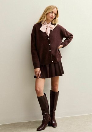 Cardigan oversize marrone con scollo a V, chiusura a bottoni e fiocco rosa chiaro al colletto, abbinato a una gonna plissettata scura e stivali marroni al ginocchio.
