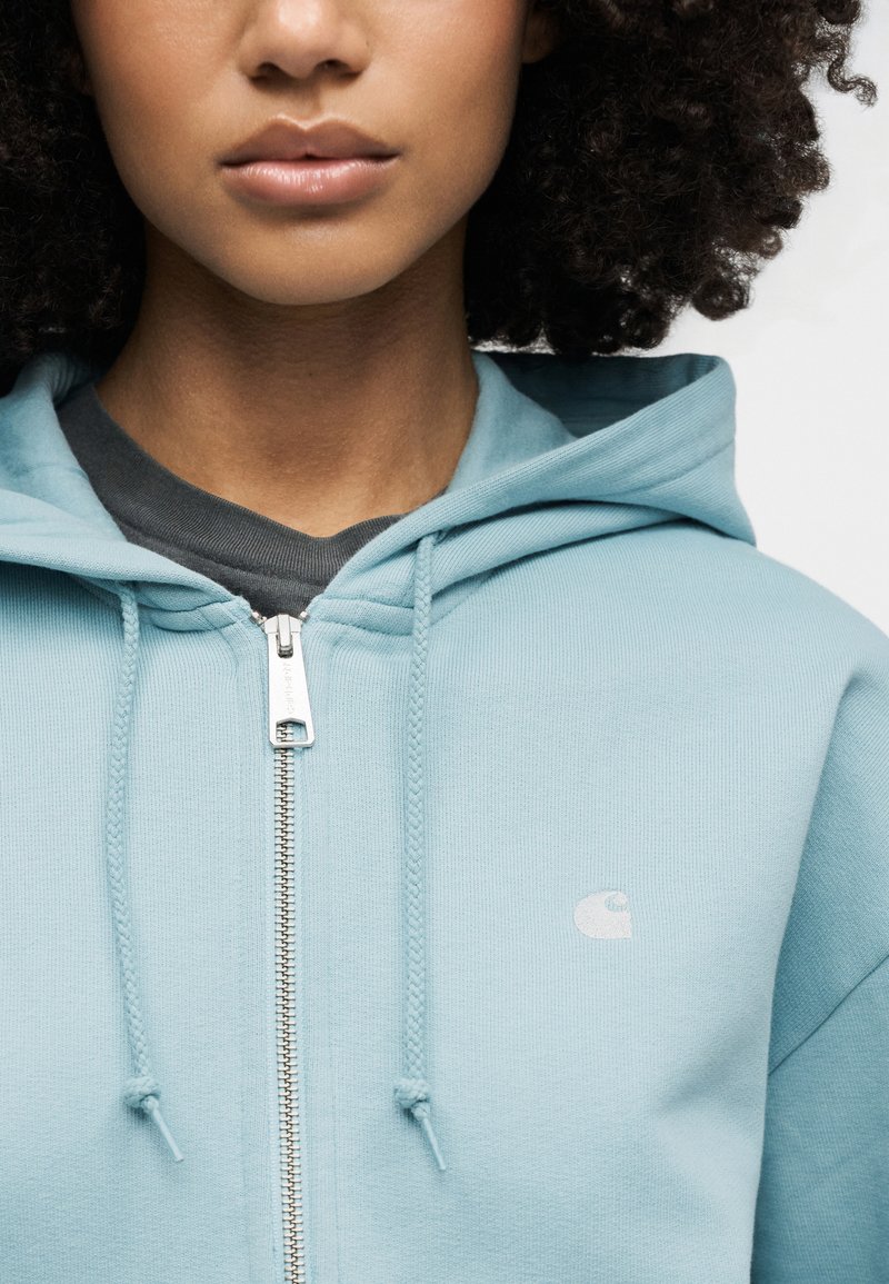 Femme aux cheveux bouclés portant un sweat à capuche zippé bleu clair par-dessus un t-shirt gris foncé, vue du cou à la poitrine.