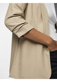 Beige sako s ohrnutými rukávy, s jemnou texturou a minimalistickým designem, nošené přes bílé tričko a černé kalhoty.