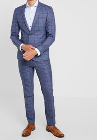 Modrý kostkovaný oblek s klopami, odpovídající kalhoty a světle modrá košile. Hnědé šněrovací boty dotvářejí celý outfit.