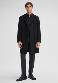 Cappotto lungo in lana nera con colletto a risvolto, tre bottoni e tasche laterali, abbinato a pantaloni scuri e scarpe di pelle nere.
