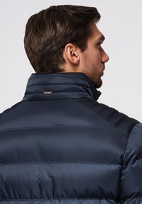 Mand med kort brunt hår iført en navy blå polstret jakke, vist fra ryggen og i sideprofil mod en ensfarvet baggrund.