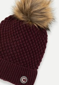 Adīts bordo cepurīte ar teksturētu rakstu, ar pūkainu pom-pomu augšpusē un apaļu logo uzlīmi uz stieptā mala.