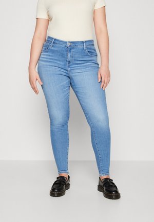 Osoba w jasnoniebieskich obcisłych dżinsach, kremowej bluzce z krótkim rękawem i czarnych mokasynach stoi na tle jednolitego jasnego tła.