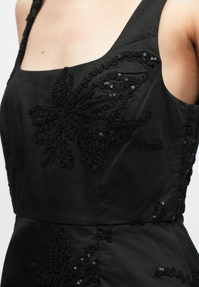 Μαύρο φόρεμα από ύφασμα με τετράγωνη λαιμόκοψη, διακοσμημένο με μαύρη λουλουδάτη κεντημένη και λεπτομέρειες από παγιέτες στον κορμό, προσφέροντας μια υφασμάτινη αίσθηση.