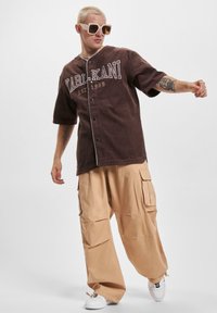 Camicia a maniche corte in velluto a coste marrone con bottoni e scritte bianche, abbinata a pantaloni cargo beige chiaro. Modella che indossa occhiali da sole oversize.