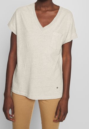Personne portant un t-shirt beige clair à col en V et manches courtes avec une poche poitrine et un pantalon beige clair sur un fond uni.