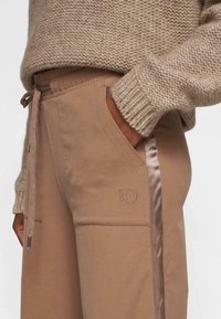 Pantalones de chándal beige con una textura suave, que presentan una franja de satén lateral y el número 10 bordado, combinados con un suéter de punto grueso en color crema.