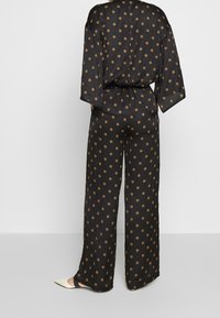 Black, silky jumpsuit featuring wide sleeves and a drawstring waist, decorated with gold circular patterns, wide-leg pants, and subtle sheen.