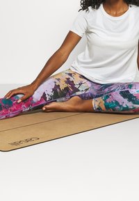 Flerfärgade leggings med abstrakta mönster i lila, rosa och turkos, kombinerade med en enkel vit t-shirt, på en beige yogamatta.