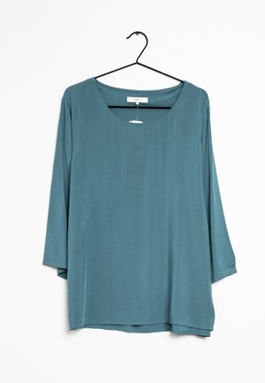 Blouse bleue à manches longues avec un col rond, suspendue sur un cintre noir devant un fond blanc.