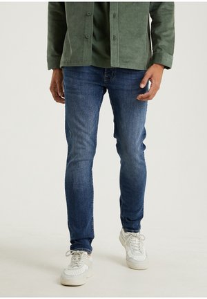 Homme portant un jean slim bleu, des baskets blanches et une veste en velours côtelé verte, debout devant un fond clair uni.