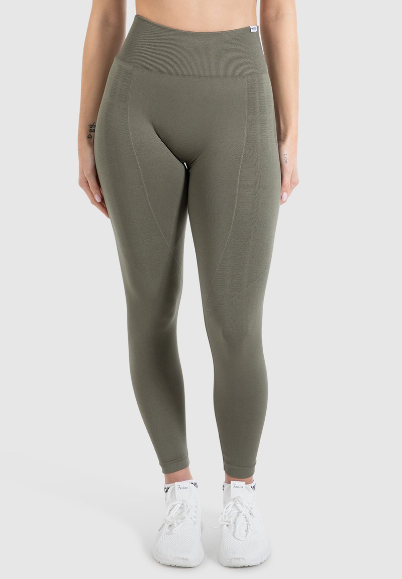 Leggings verde oliva a vita alta con una vita a costine e pannelli texturizzati. Indossati con scarpe da ginnastica bianche. Tessuto liscio e elasticizzato.