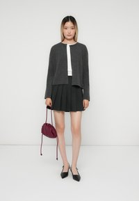 Femme se tient contre un mur blanc, portant un cardigan gris, un haut blanc, une jupe plissée noire, des chaussures à talons noirs, tenant un petit sac bordeaux.