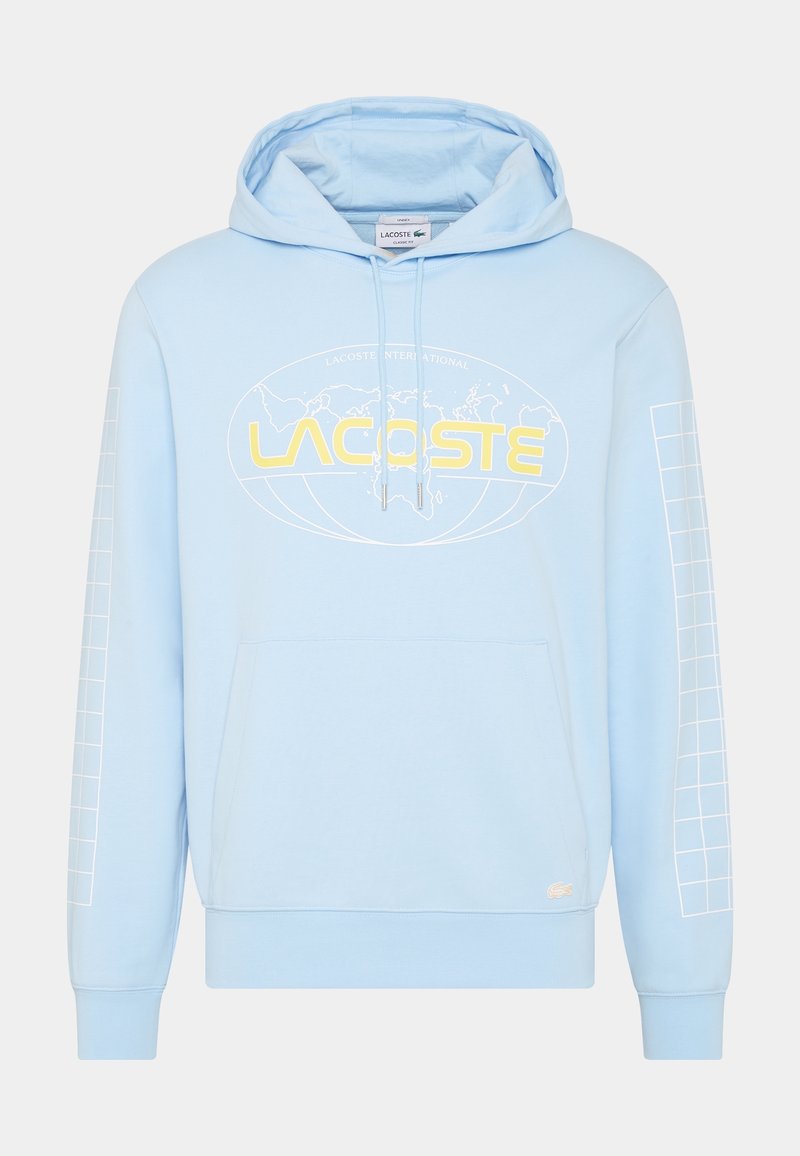 Sweat à capuche bleu clair avec poche avant, texte jaune "LACOSTE" sur un graphisme de carte du monde, et lignes de quadrillage blanches sur les manches.
