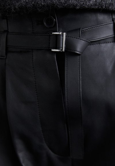 Μαύρα δερμάτινα παντελόνια με κομψό σχέδιο, ζώνη με ασημένια αγκράφα και λεπτομερή ραφή κατά μήκος των ραφών. Φαν�ερή λεία υφή.
