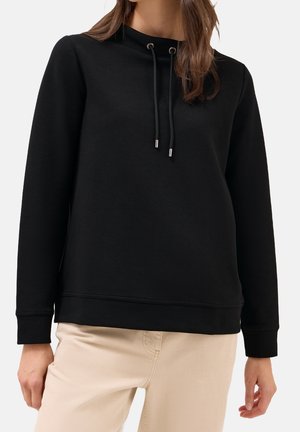 Femme portant un pull noir à manches longues avec cordons et un pantalon beige sur un fond clair uni.