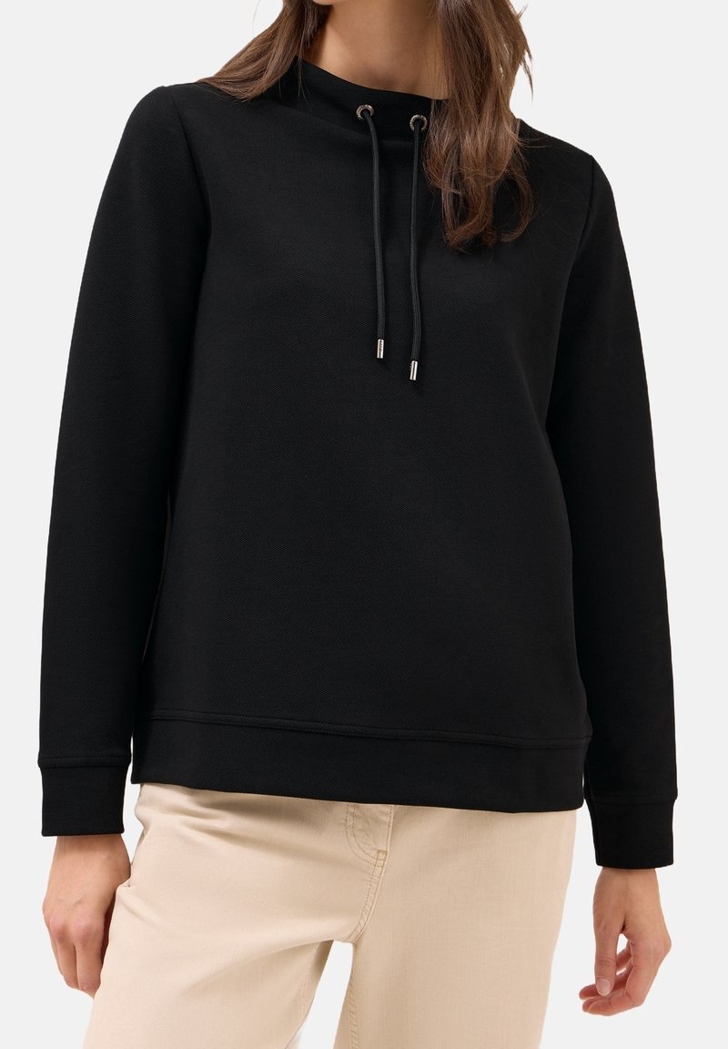 Femme portant un pull noir à manches longues avec cordons et un pantalon beige sur un fond clair uni.