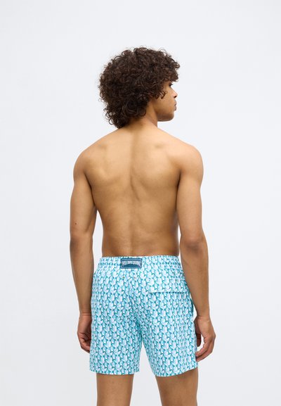 Muž s kučeravými vlasmi, ktorý má na sebe modro-biele plavky s vzorom, otočený chrbtom k jednofarebnému svetlému pozadiu.