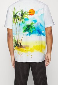 Camisa blanca de manga corta con un diseño de paisaje en acuarela que incluye palmeras, un sol y una montaña en tonos azules y amarillos.