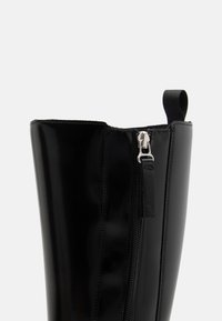 Sort blank glossy knæhøj støvle med sidelås og trækflap mod en ensartet lys baggrund.