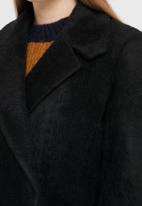 Abrigo negro con un tejido texturizado y peludo. Presenta un cuello ancho y un corte holgado, costuras visibles y una apariencia suave.