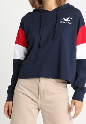 Mörkblå huvtröja med röda och vita horisontella ränder på ärmarna; har en frontficka i kängurustil och en logotyp på bröstet; material i bomullsblandning.