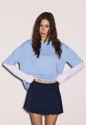 Jeune femme aux longs cheveux ondulés portant une chemise oversize bleu clair, des manches blanches en dessous, et une jupe plissée bleu foncé, debout contre un mur uni.