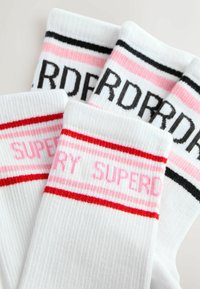 Valged puuvillased sokid, millel on soonikkoes, roosa, punase ja musta triibulisusega ning "SUPERDRY" tekst roosa ja musta värviga.