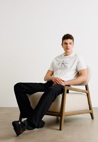 T-shirt bianca con logo grigio, pantaloni neri e scarpe nere. Seduto su una sedia rivestita beige chiaro con struttura in legno.
